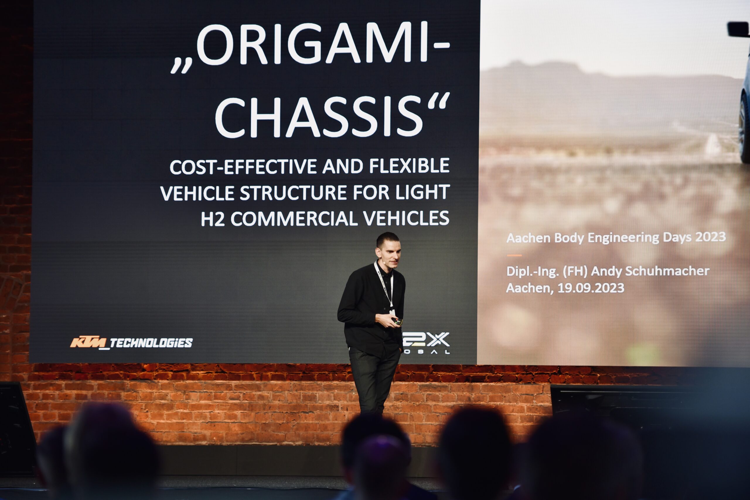 Man doing a presentation on stage. Large screen behind saying Origami Chassis.